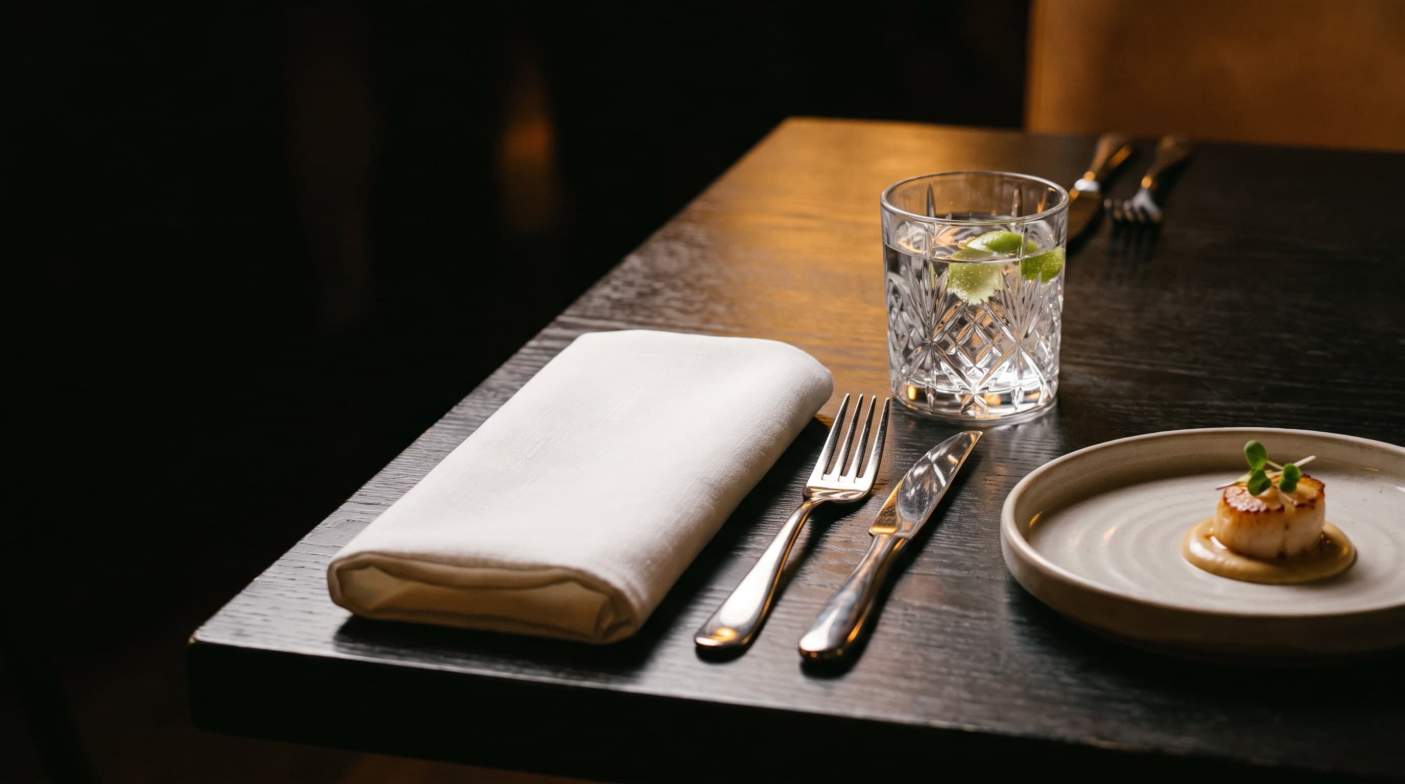 Weisse Serviette, Silberbesteck, Kristallglas und ein Amuse-Bouche auf einem dunklen Tisch im Spotlight